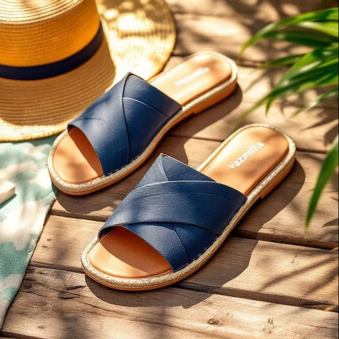 Blue sandals with tan soles on a wooden surface with a straw hat and plant in the background.